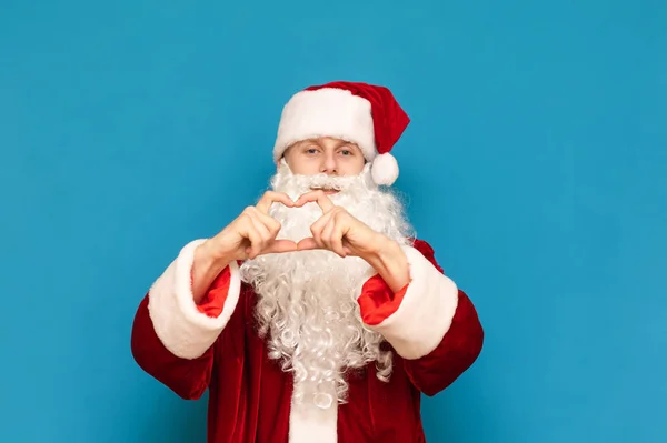 Przystojny młody Święty Mikołaj pokazuje gest serca rękami i patrzy w kamerę z pozytywną twarzą odizolowaną na niebieskim tle. Mikołaj cię kocha. Boże Narodzenie i Nowy Rok. — Zdjęcie stockowe