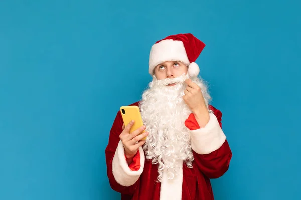 Porträtt av en pensiv jultomte med hjälp av en smartphone, vrida sin mustasch och tittar tankeväckande uppåt mot en blå bakgrund. Killen i tomtedräkt och smartphone i handen är isolerad. Nytt år — Stockfoto