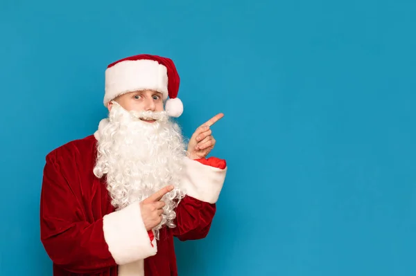 Lustiger Typ im Weihnachtsmann-Anzug isoliert auf blauem Hintergrund, zeigt auf den leeren Raum, schaut in die Kamera und lächelt. Positiver Weihnachtsmann für Weihnachten und Copyspace — Stockfoto