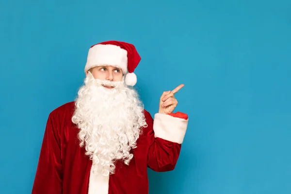 Das Porträt eines Mannes im Weihnachtsmannanzug steht auf blauem Hintergrund und zeigt seinen Finger zur Seite auf einem leeren Sitz. junge Weihnachtsmann zeigt Hand auf Copyspace. Weihnachtsfasching. — Stockfoto