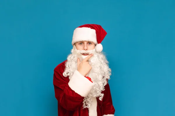 Portret zabawnej, zamyślonej nastolatki w stroju Świętego Mikołaja, patrzącej uważnie w kamerę, odizolowanej na niebieskim tle. Młody Mikołaj w kostiumie patrzy w aparacie i myśli.Boże Narodzenie i Nowy Rok — Zdjęcie stockowe