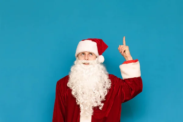 Cute młody Święty Mikołaj odizolowany na niebieskim tle, patrząc i wskazując palcem w puste miejsce. Wesoły facet Mikołaja wskazuje palcem na Copyspace. Koncepcja przyjęcia świątecznego. — Zdjęcie stockowe