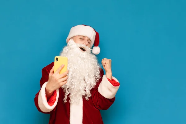 Unge jultomten vinnare jublar i seger med smartphone i handen på blå bakgrund, ser in i kameran med glada ansikte. Framgångsrik spelare spelar mobilspel på smartphone. Nytt år och jul. — Stockfoto