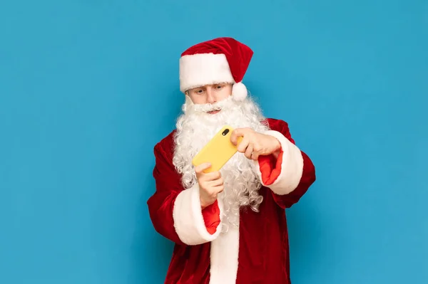 Chico en traje de carnaval de Santa Claus jugando juegos móviles en el teléfono inteligente aislado sobre fondo azul. El sitio joven juega juegos en línea y teléfonos inteligentes, mira atentamente a la pantalla del teléfono inteligente . — Foto de Stock