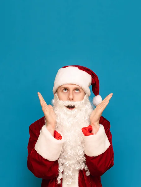 Retrato de un tipo asustado en un traje de Santa Claus sobre un fondo azul, sorprendido con las manos levantadas para mirar hacia arriba en un lugar en blanco. Sorprendido joven Santa mirando el espacio de copia. Vertical — Foto de Stock