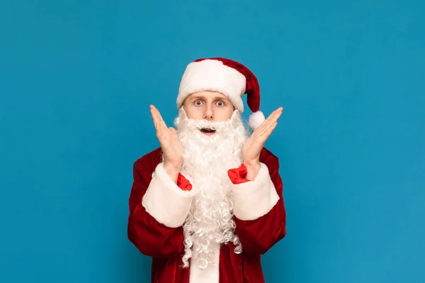 Retrato de un joven Santa Claus asustado sobre un fondo azul, se ve impactado cara a cámara y levantó las manos para enfrentarse con miedo. Aislado. Copiar espacio — Foto de Stock