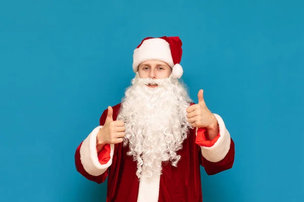 Fröhliche junge Weihnachtsmänner im Anzug stehen vor blauem Wandhintergrund und zeigen Daumen hoch, schauen in die Kamera und lächeln. Santee mag es. Weihnachts- und Neujahrskonzept. — Stockfoto