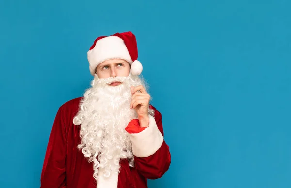 Nahaufnahme Porträt eines nachdenklichen Weihnachtsmannes auf blauem Hintergrund, der in einen leeren Raum blickt und denkt. Junger Kerl im Weihnachtsmannanzug isoliert auf blauem Hintergrund. Weihnachtskonzept. — Stockfoto