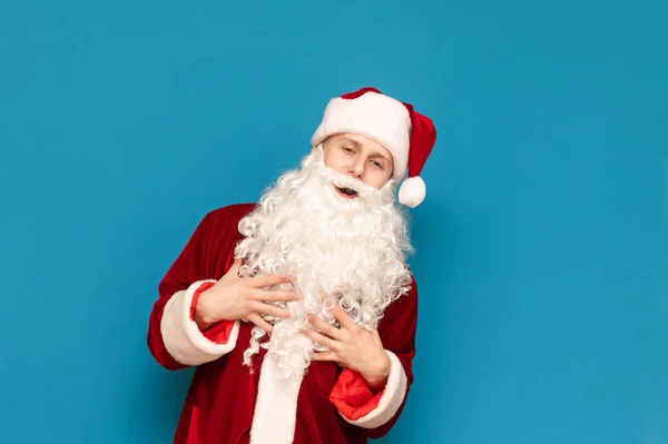 Positiv ung man i Santa kostym står på blå bakgrund, ler och tittar på kameran. Ung glad tomte skrattar, isolerad. Julkoncept. — Stockfoto