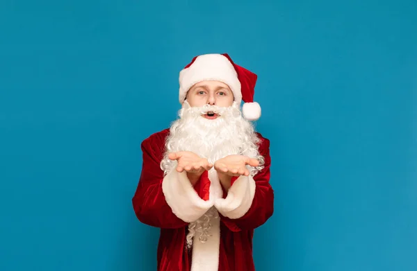 Portrait de jeune homme positif en costume de Père Noël regardant dans la caméra et envoyant un baiser sur un fond bleu. Père Noël tient la paume près du visage et souffle dans la caméra . — Photo