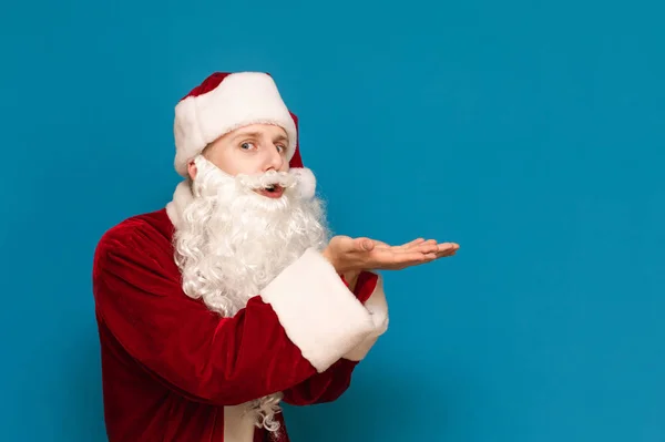 Retrato de un divertido Santa Claus joven soplando sus manos a un lado y mirando a la cámara con una cara sorprendida. Chico alegre en traje de santa golpes en la palma de la mano a un lado. Concepto de Navidad . — Foto de Stock