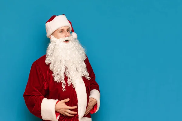Rolig ung man i jultomten kostym och stor mage poserar på kameran mot blå bakgrund. Roliga unga tomten håller hennes mage och tittar in i kameran. — Stockfoto