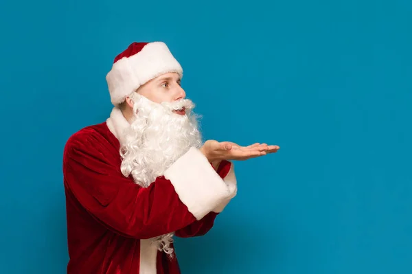 Jeune Père Noël regarde loin et souffle à l'espace de copie. Le type en costume Père Noël montre ses mains sur le côté et souffle ses mains sur l'espace de copie. Père Noël montre air baiser . — Photo