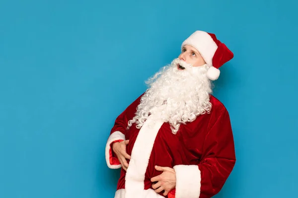 Divertido joven Santa con barba, sombrero y vientre grande aislado sobre fondo azul, mirando hacia otro lado. Un adolescente se vistió con un disfraz de Papá Noel y posa para la cámara. Concepto de Navidad . — Foto de Stock