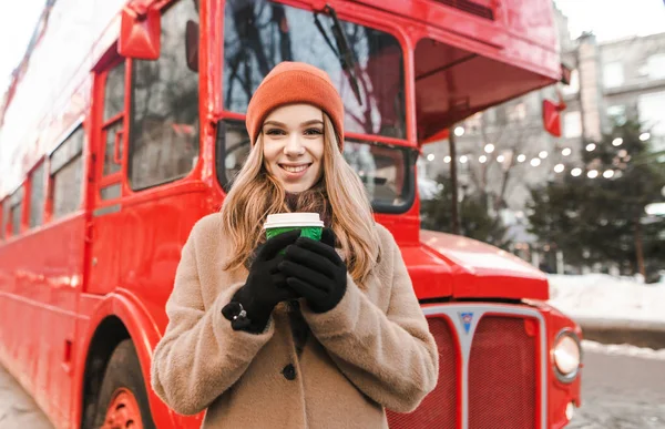 Πορτρέτο ενός χαμογελαστού κοριτσιού με χειμωνιάτικα ρούχα στέκεται στο δρόμο με ένα φλιτζάνι καφέ στα χέρια της σε φόντο κόκκινου λεωφορείου, κοιτάζει στην κάμερα και χαμογελά. Χαριτωμένη έφηβη σε μια βόλτα σε μια χειμωνιάτικη μέρα. — Φωτογραφία Αρχείου