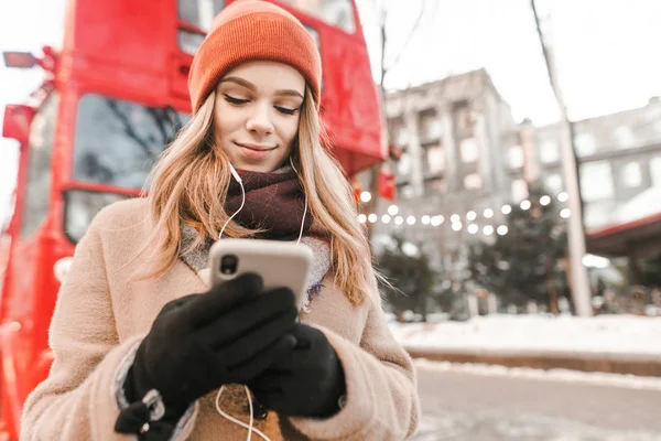 Χαριτωμένο κορίτσι σε ζεστά ρούχα και γάντια στέκεται στο δρόμο και χρησιμοποιεί ένα smartphone σε ένα αστικό φόντο το χειμώνα, ακούγοντας μουσική στα ακουστικά, Χειμώνας με τα πόδια ενός όμορφου κοριτσιού με μουσική — Φωτογραφία Αρχείου