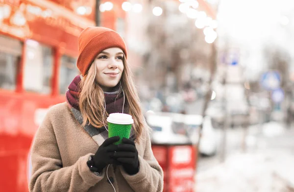 Χειμερινό πορτρέτο δρόμου ενός χαριτωμένου κοριτσιού με ζεστά ρούχα στο δρόμο, στέκεται με ένα φλιτζάνι ζεστό ποτό στα χέρια της, κοιτάζοντας μακριά με ένα χαμόγελο. Χριστουγεννιάτικη φωτογραφία του θετικού κοριτσιού στο δρόμο. — Φωτογραφία Αρχείου