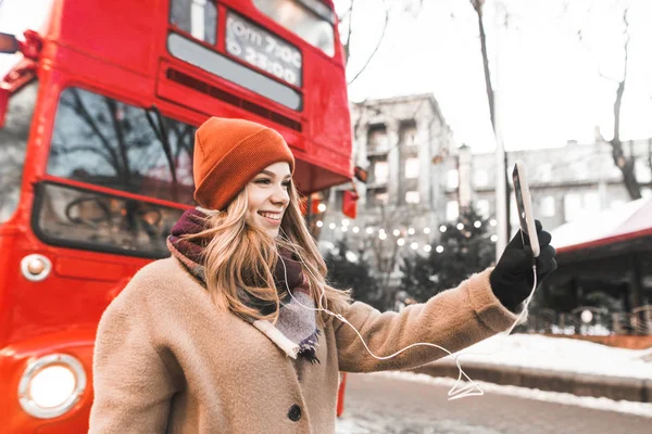 Όμορφη κοπέλα με ζεστά ρούχα στέκεται σε ένα δρόμο το χειμώνα με ακουστικά και παίρνει μια selfie σε ένα smartphone στο χέρι της. Ελκυστικό χαμογελαστό κορίτσι κάνει τη φωτογραφία της στην κάμερα smartphone. — Φωτογραφία Αρχείου