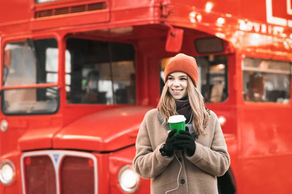 Πορτρέτο του ευτυχισμένου κοριτσιού με ζεστά ρούχα σε μια βόλτα σε μια κρύα μέρα του χειμώνα, στέκεται σε ένα φόντο ενός κόκκινου λεωφορείου με ένα φλιτζάνι καφέ στα χέρια της, κοιτάζοντας μακριά. Χριστουγεννιάτικη ιδέα. — Φωτογραφία Αρχείου