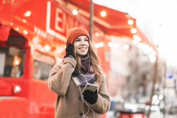 Ευτυχισμένο κορίτσι με τα πόδια το χειμώνα για τα Χριστούγεννα διακοσμημένο δρόμο με smartphone στο χέρι, ακούγοντας μουσική στα ακουστικά, κοιτάζοντας μακριά και χαμογελώντας. Χριστουγεννιάτικο πορτρέτο δρόμου. — Φωτογραφία Αρχείου