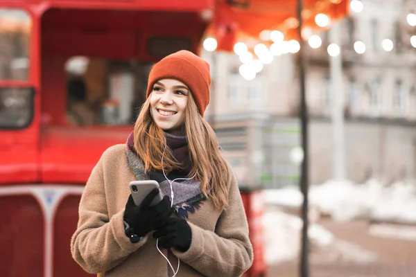 Χαμογελαστή κοπέλα στέκεται στο δρόμο Χριστούγεννα διακοσμημένα με smartphone στο χέρι, ακούει μουσική, κοιτάζει μακριά. Οδός πορτρέτο χειμώνα ενός χαριτωμένο κορίτσι τα Χριστούγεννα στο δρόμο. — Φωτογραφία Αρχείου
