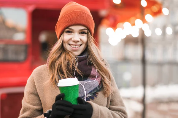 Κοντινό πορτρέτο του χαμογελαστού κοριτσιού με ακουστικά και φλιτζάνι καφέ στα χέρια, φορώντας ζεστά ρούχα, στέκεται το χειμώνα σε εξωτερικούς χώρους φωτεινό φόντο κοιτάζοντας στην κάμερα.Χριστουγεννιάτικο πορτρέτο ενός κοριτσιού — Φωτογραφία Αρχείου