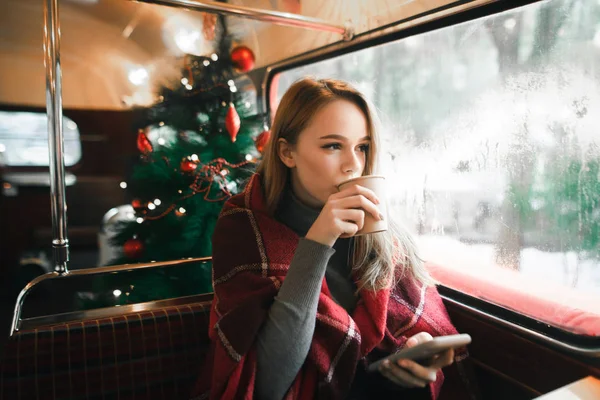 Πορτρέτο ενός χαριτωμένο κορίτσι κάθεται τα Χριστούγεννα σε ένα ζεστό καφέ, πίνοντας καφέ, κοιτάζοντας έξω από το παράθυρο και κρατώντας ένα smartphone στο χέρι της.Πορτρέτο όμορφο κορίτσι στο καφέ στο φόντο χριστουγεννιάτικο δέντρο — Φωτογραφία Αρχείου