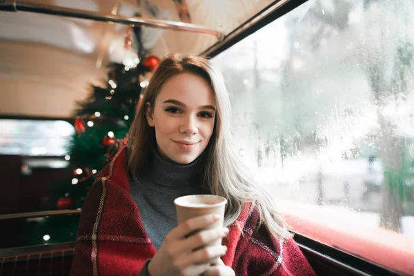 Belle fille assise dans un café confortable près de la fenêtre avec une tasse de café dans sa main regarde dans la caméra sur le fond de l'arbre de Noël. Belle fille boire du café dans le café et à Chri — Photo