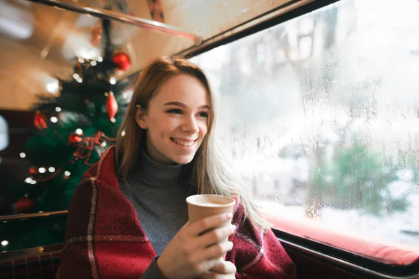 Happy girl sitting at Christmas in a cozy cafe with Christmas tree, holds a cup of coffee in her hands, looks out the window. Winter portrait of happy girl warming up in coffee drink. — Stock Photo, Image