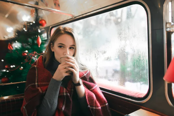 Cute girl in plaid on the background of the Christmas tree sitting in a cozy cafe and warmed by a warm drink. Cute lady drinking coffee in a coffee shop and looking away. — Stock Photo, Image