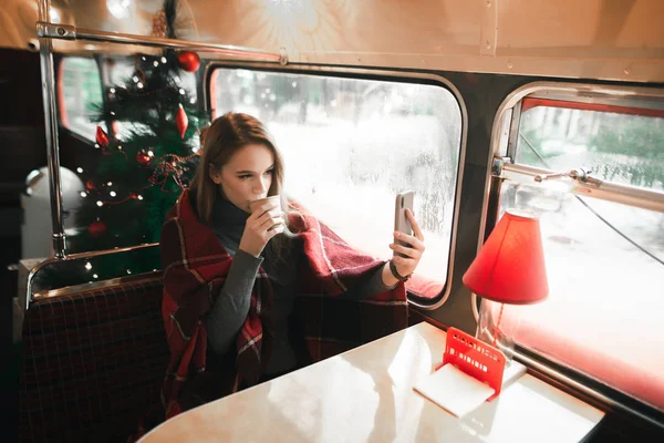 Nettes Mädchen, das im Winter in einem gemütlichen Café sitzt, Kaffee trinkt und vor dem Hintergrund des Weihnachtsbaums Selfies macht. Schöne Dame im Karo macht Selfie in weihnachtlich dekoriertem Café — Stockfoto