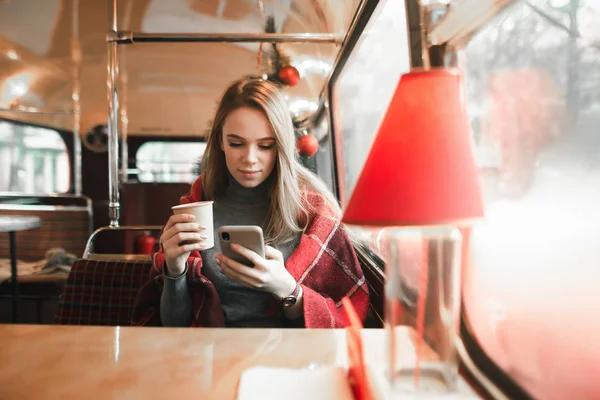 Симпатичная девушка сидит в уютном кафе на фоне елки с чашкой кофе в руках и концентрируется на использовании смартфона. Девушка сидит в кафе на Рождество . — стоковое фото