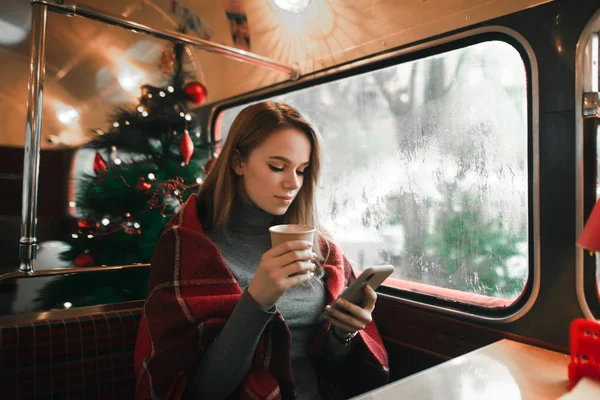 Chica bonita sentada en un acogedor café de Navidad con una taza de café en la mano, utiliza un teléfono inteligente, se ve en la pantalla con una cara seria. Linda dama pasa el día de invierno en un acogedor café con una bebida caliente . — Foto de Stock