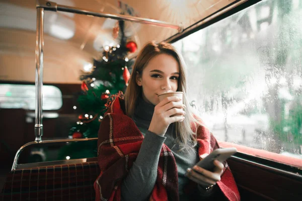 Όμορφη κοπέλα σε ένα ζεστό καφέ στο φόντο ενός χριστουγεννιάτικου δέντρου με ένα smartphone στο χέρι της, κοιτάζει στην κάμερα με ένα σοβαρό πρόσωπο και πίνει καφέ. Κορίτσι που κάθεται στο καφέ σε ένα κρύο χειμώνα da — Φωτογραφία Αρχείου