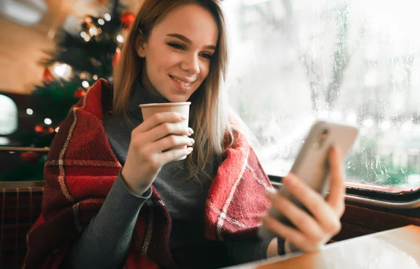 Girl sitting in cozy cafe near window and making photo on smartphone with cup of coffee, cute girl taking selfie on Christmas tree background in coffee shop.Lady uses smartphone in winter in cafe