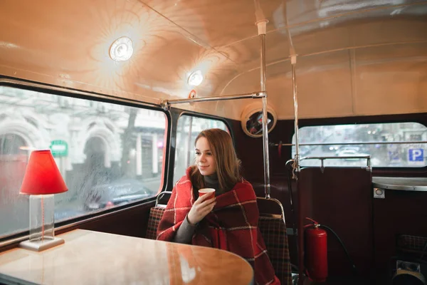 Cute girl spends a cool winter day in a cozy cafe with a cup of coffee, looks out the window and smiles. Beautiful girl basking in plaid and drinking coffee. — ストック写真