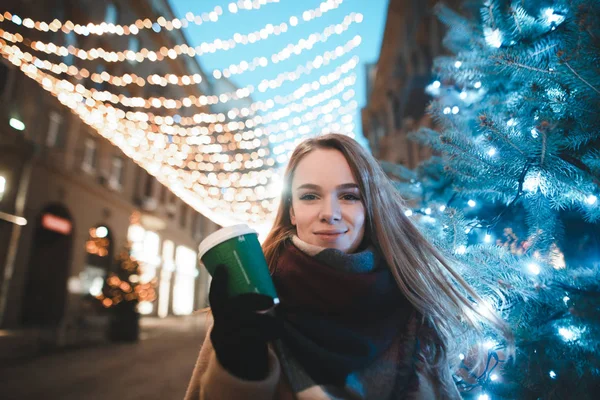 Πορτρέτο του όμορφου κοριτσιού με ζεστά ρούχα και μαντήλι σε μια βόλτα σε ένα κρύο βράδυ του χειμώνα, στέκεται κοντά σε ένα χριστουγεννιάτικο δέντρο με ένα λαιμό στο δρόμο και κρατά ένα φλιτζάνι καφέ στο χέρι της, κοιτάζει στην κάμερα — Φωτογραφία Αρχείου