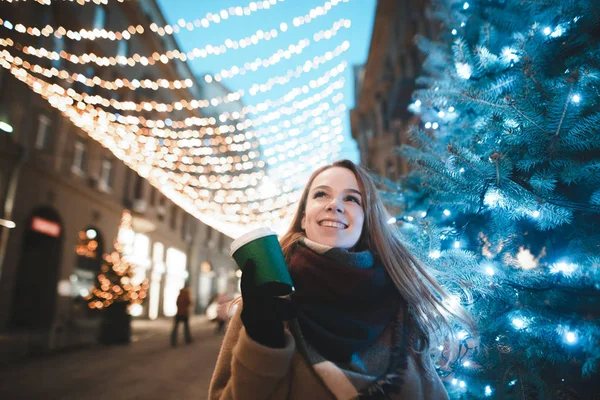 Happy girl walking in the evening on the street decorated for Christmas street, stands near a Christmas tree with a cup of coffee, looks up and smiles. Walk in the Christmas — ストック写真