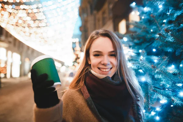 Κοντινό χριστουγεννιάτικο πορτρέτο ενός χαμογελαστού κοριτσιού στο δρόμο κοντά σε ένα χριστουγεννιάτικο δέντρο στο φόντο ενός δρόμου διακοσμημένου για τα Χριστούγεννα, κοιτάζοντας την κάμερα και χαμογελώντας. Νέο Έτος έννοια. — Φωτογραφία Αρχείου