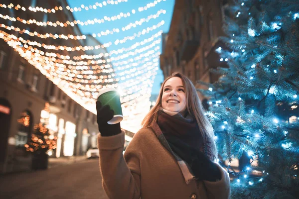 Joyful girl walks down the street in the evening with Christmas lights and a Christmas tree, holds a cup of coffee, looks up and smiles. Christmas evening portrait of a girl. New Year concept. — ストック写真