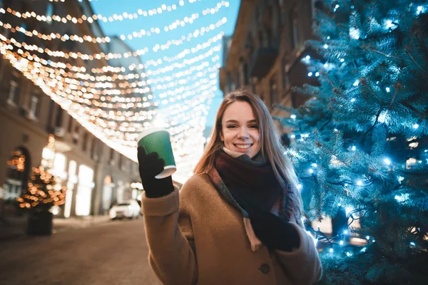 Ένα χαρούμενο κορίτσι στέκεται σε ένα χριστουγεννιάτικο δρόμο με ένα φλιτζάνι καφέ στα χέρια της, κοιτάζει στην κάμερα και χαμογελάει. Πορτρέτο ενός χαριτωμένο χαμογελαστό κορίτσι σε εξωτερικούς χώρους κοντά σε ένα χριστουγεννιάτικο δέντρο με φώτα. — Φωτογραφία Αρχείου
