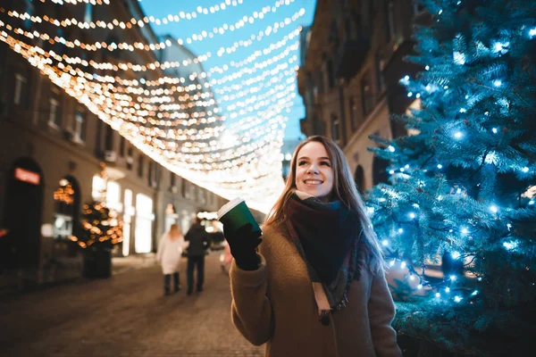 Street photo of happy girl at christmas evening near standing near fir tree on decorated light street with cup of coffee in her hands and smiling. Portrait of a cute lady on New Year's night. — ストック写真