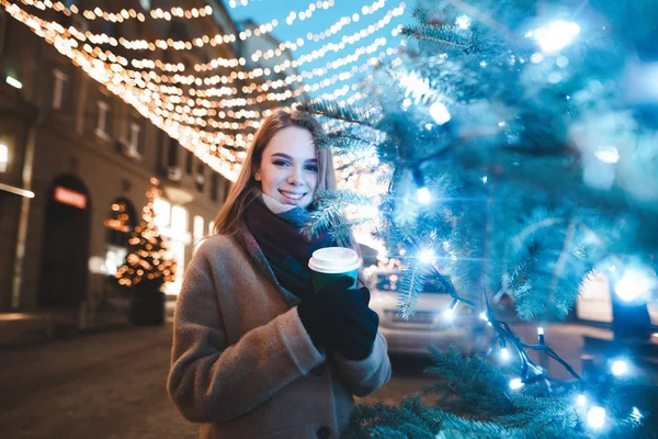 Happy lady walking on the street on a winter evening at Christmas, looking at the camera and drinking coffee, posing near a Christmas tree. New Year and Christmas concept. Christmas street scenery. — ストック写真