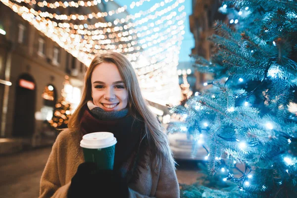 Christmas portrait of a beautiful girl in warm clothing and scarf stands on the street decorated for New Year, looks at the camera and rejoices, holds a cup of coffee. Walk for Christmas. — Stock Photo, Image