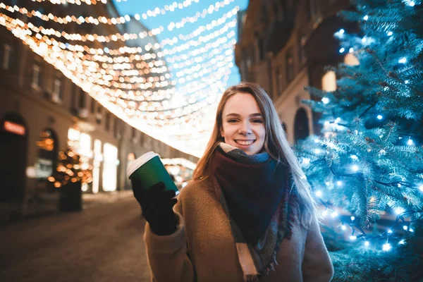 Ένα χαμογελαστό κορίτσι με ζεστά ρούχα στέκεται σε ένα χριστουγεννιάτικο δρόμο κοντά σε ένα χριστουγεννιάτικο δέντρο με ένα φλιτζάνι καφέ στο χέρι της, κοιτάζει στην κάμερα και γελάει. Χριστούγεννα και Πρωτοχρονιά cocnept. — Φωτογραφία Αρχείου