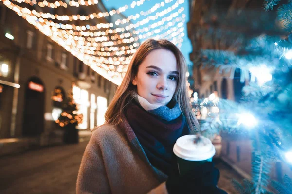 Menina em um cachecol e casaco fica perto de uma árvore de Natal na rua com uma xícara de café na mão e olha para a câmera. Rua retrato de Natal de bela senhora no fundo da rua com lanternas . — Fotografia de Stock