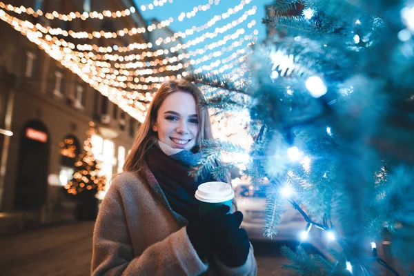 Retrato de uma menina bonita contra o fundo da rua decorações de Natal, beber café e olhar para a câmera com um positivo. Menina sorrindo andando pela rua para o Natal — Fotografia de Stock