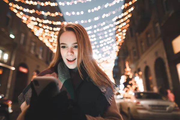 Retrato de menina surpresa à noite na rua decorada com uma grinalda, olha para a tela de um smartphone. Menina chocada usa smartphone à noite na rua atmosférica . — Fotografia de Stock