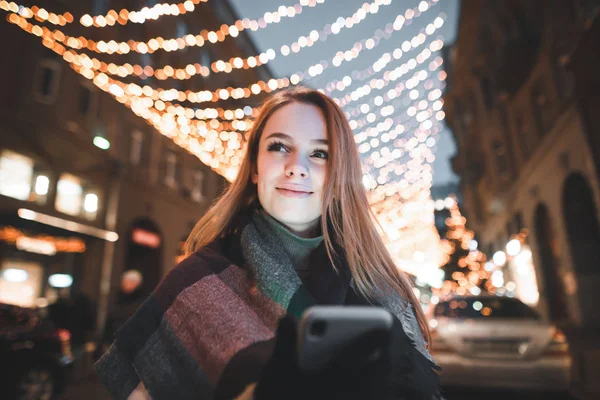 Close-up de menina sorridente com smartphone em mãos no fundo da rua com luzes de Natal, olhando para o lado e sorrindo. Noite retrato de uma menina bonito com um telefone na mão . — Fotografia de Stock