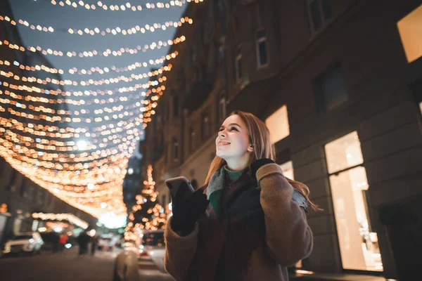 Satisfied girl stands in the street with Christmas lights, holds a smartphone in her hand, looks up and smiles. Joyful girl uses smartphone and smiles. Christmas portrait. — ストック写真